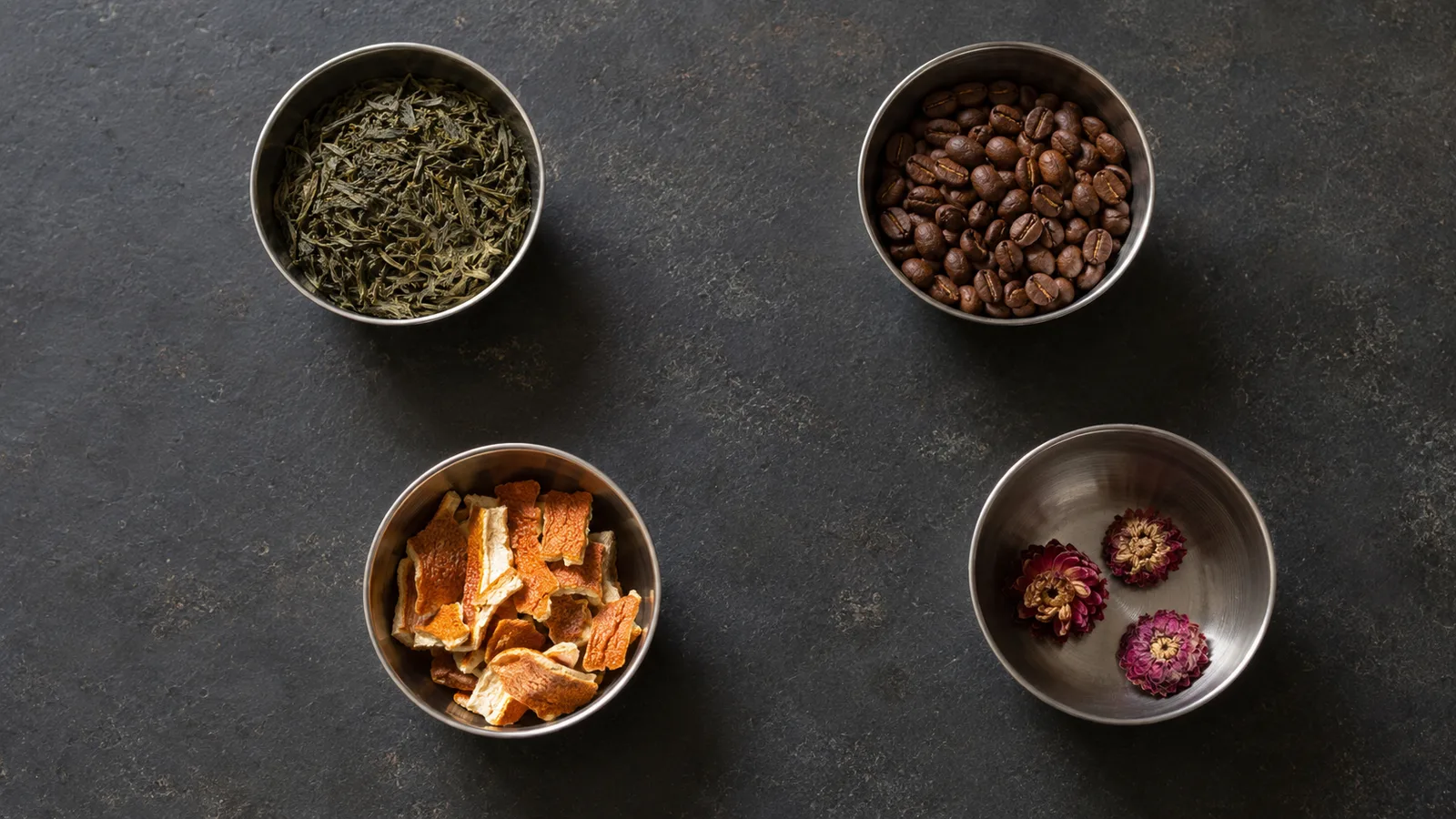 Loose green tea leaves, coffee beans, dried orange peel and dried flowers in small steel bowls